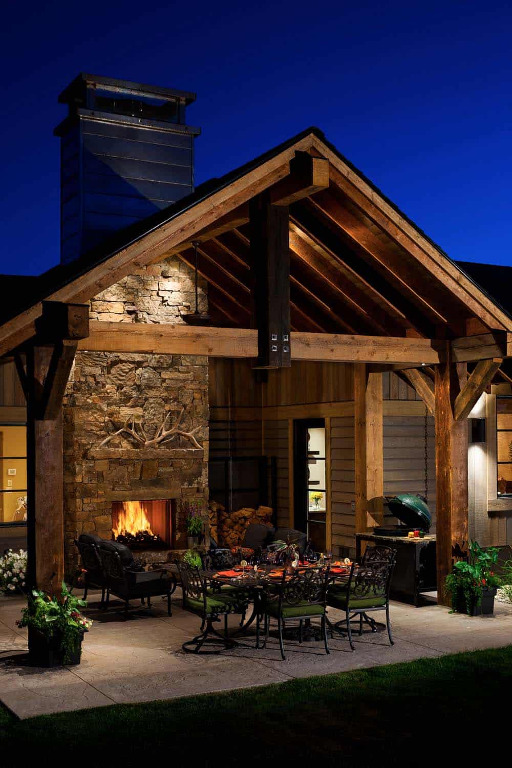 rustic mountain house exterior with a covered backyard patio and a fireplace at dusk