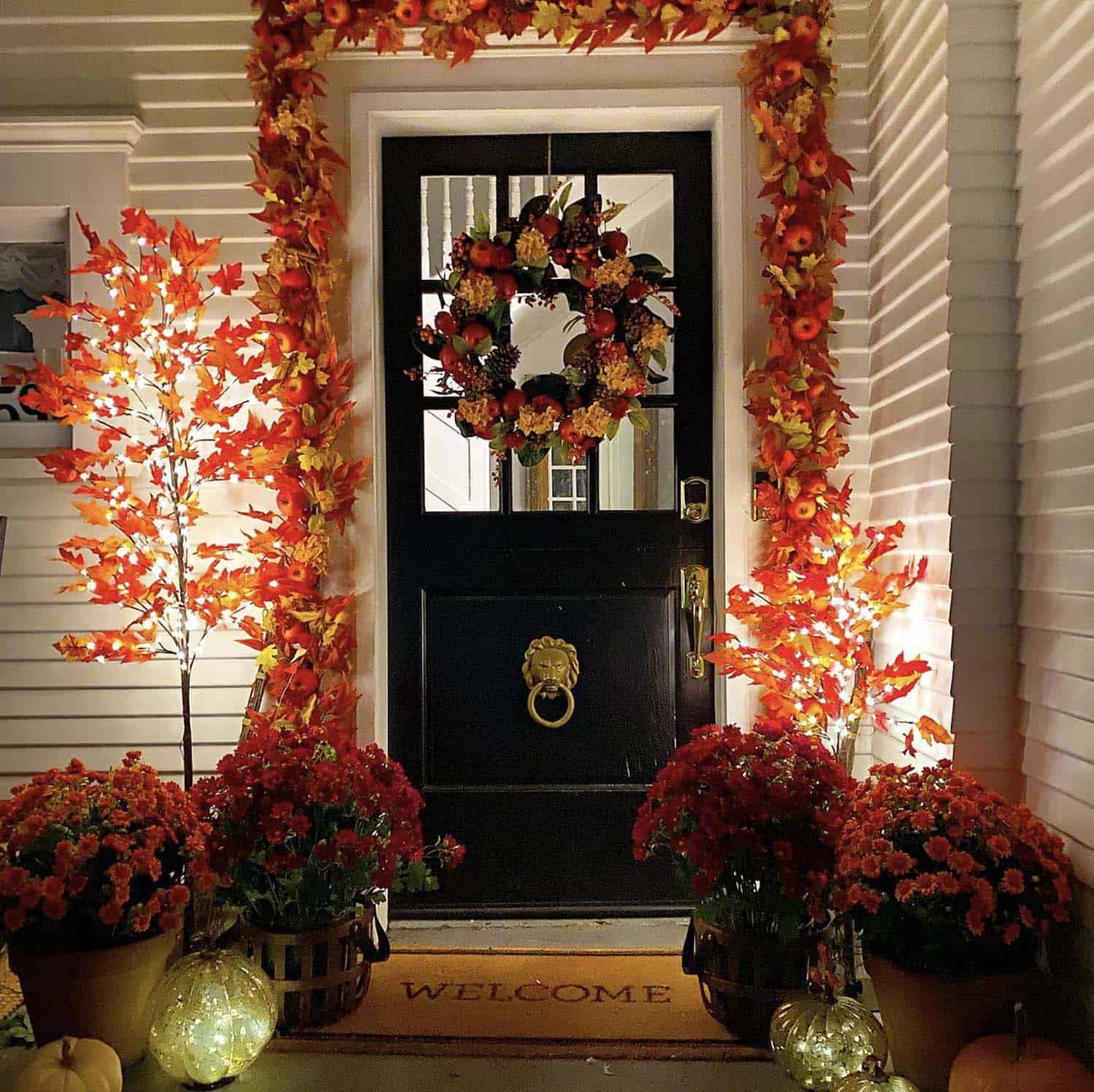 fall front porch decor at night with a garland, wreath, mums and lighted pumpkins