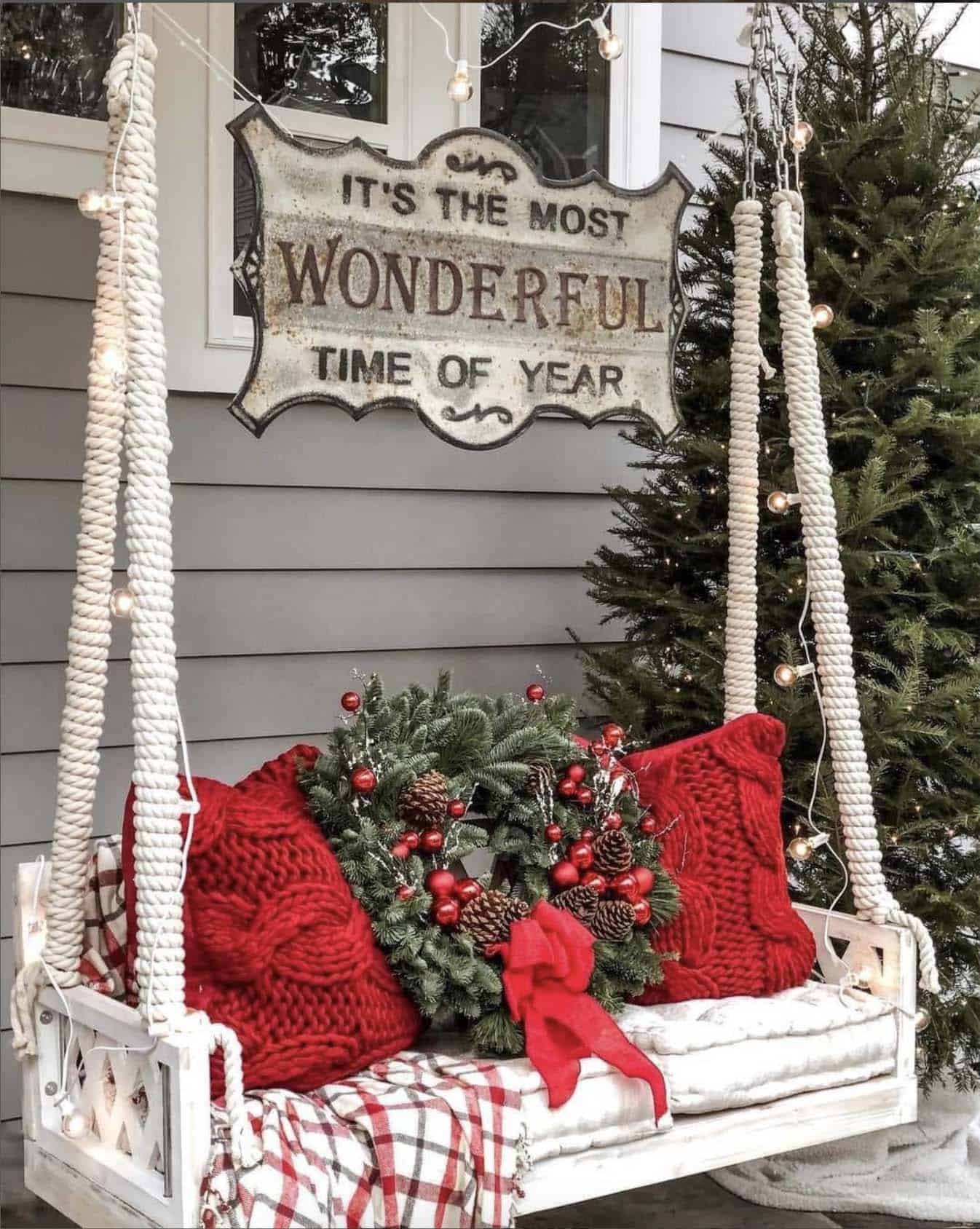 cozy porch swing with red pillows, a wreath, a sign and a Christmas tree
