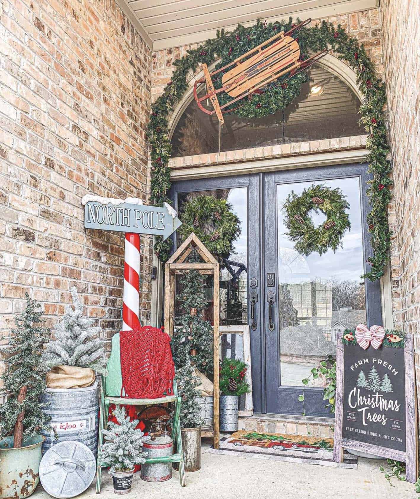 farmhouse front porch with Christmas trees, holiday signs, garland, and front door wreaths